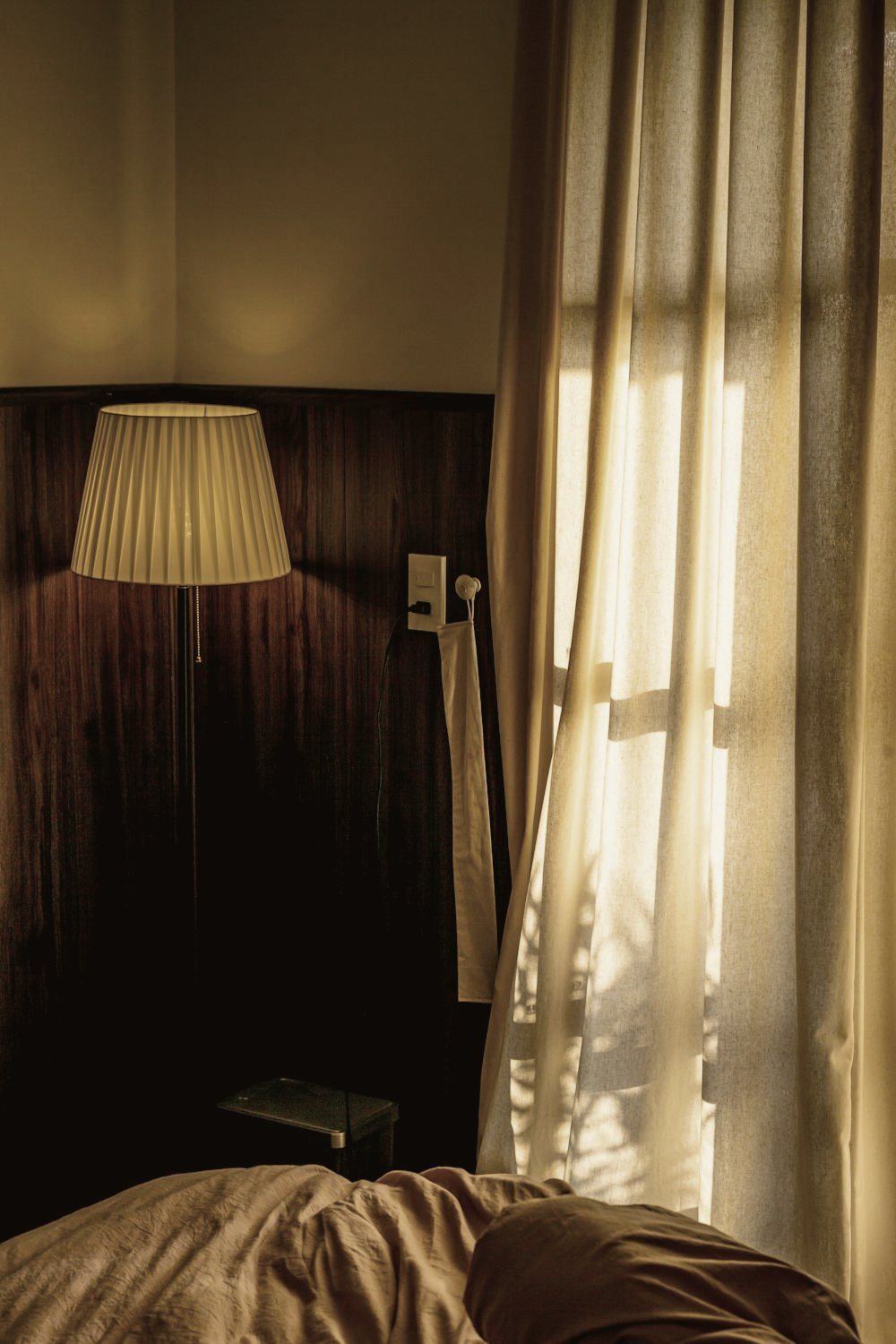 A calm bedroom corner at golden hour: a floor lamp, sheer linen curtains, a phone charging on the bedside table and a socket beside the bed